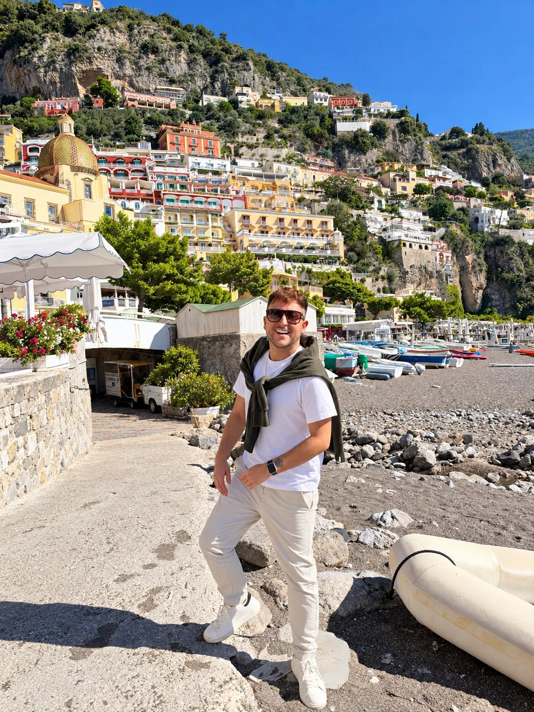 Luke Brennan smiling in sunglasses on a pebble beach in Positano, with colourful hillside buildings behind him